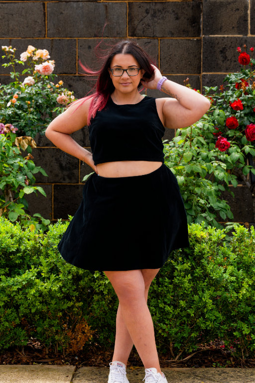 A woman modelling a black velvet sleeveless crop top and mini skirt, standing in front of a bluestone wall and rose bushes.